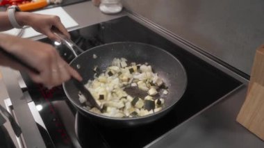 Bir kadın, indüksiyon sobasında kabak ve soğan doğruyor. Sebzeler siyah bir spatulayla karıştırılır ve yumuşayana kadar pişirilir..