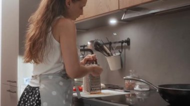 Puantiyeli önlük giyen bir kadın, havucu rendeleyerek ızgara yapar. Günlük rutininin bir parçası olarak yemek hazırlıyor. Etrafı mutfak aletleri ve dilimlenmiş sebzelerle çevrili..