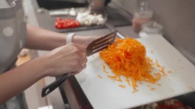 Bir kadın indüksiyon sobasındaki tavaya rendelenmiş havuç ekliyor. Havuçlar kabak, dolmalık biber ve soğanla karıştırılır. Sağlıklı bir yemek hazırlığı için sotelenir.