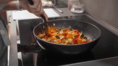 Bir kadın indüksiyon sobasında rendelenmiş havuç, kabak, soğan ve dolmalık biberi yapışık olmayan bir tavada spatulayla karıştırıyor. Renkli sebzeler sağlıklı bir yemek için eşit olarak birleştirilir..