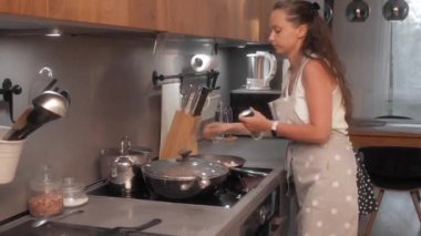 Önlüklü bir kadın mutfakta yemek pişirirken biber değirmeni kullanıyor. Fırının başında duruyor, fırının üstünde tencere ve tavalarla yemek hazırlıyor..