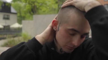 Dövmeli genç bir adam dışarıda oturuyor, stres ve endişelerini dile getiriyor. Adam kafasını elinde tutuyor, duygusal mücadele, zihinsel yorgunluk ve iç karışıklığı gösteriyor. Yüz ifadelerine yakın çekim.