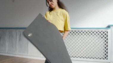 Kadın yoga minderini açar, üstünde durur, ve kollarını yukarı doğru uzatır, yoga ya da form rutinine hazırlanır. Kapalı alanda egzersiz hazırlama, esneme ve esneklik egzersizleri..