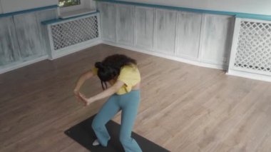 Bir kadın stüdyoda yoga minderinin üzerinde dururken kollarını ve vücudunu uzatıyor. Vücudunun esnekliğini göstermek için öne doğru eğilip eğiliyor. Yoga, esneme ve zindelik içeriği için uygundur.