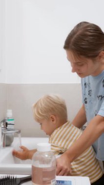 Küçük çocuk banyo lavabosunda ellerini ve yüzünü akan suyun altında yıkıyor. Anne sabırla oğluna günlük hijyen rutinini nasıl yapacağını öğretiyor. Anne bakımı ve aile hayatı..