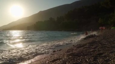 Turistler gün batımında güneşin altında denizde yüzerler. Hırvatistan 'ın Brac adasındaki Zlatni Rat plajının kumlu çakıl taşı sahilinde deniz dalgalarının tepeleri süzülüp beyaz köpüğe dönüşüyor.