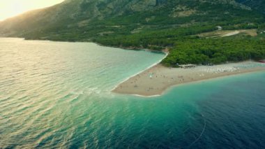 Gün batımında Zlatni Rat plajı. İnsanlar denizde yüzer, dalgalarda sallanır ve gün batımını seyreder, sahilde güneşlenir ve dinlenir. Hırvatistan 'ın Adriyatik kıyısında turizm ve plaj eğlencesi.