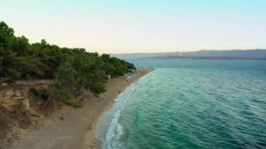 Gün batımında Zlatni Rat plajı. İnsanlar denizde yüzer, dalgalarda sallanır ve gün batımını seyreder, sahilde güneşlenir ve dinlenir. Hırvatistan 'ın Adriyatik kıyısında turizm ve plaj eğlencesi.