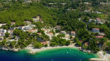Sahildeki dağ yamacında oteller ve evler. Turistler plajda güneşlenir, denizde yüzer ve kürek çekerler. Hırvatistan 'ın pitoresk Adriyatik kıyısında bir sahil turizm kasabası.