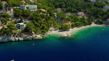 Sahildeki dağ yamacında oteller ve evler. Turistler plajda güneşlenir, denizde yüzer ve kürek çekerler. Hırvatistan 'ın pitoresk Adriyatik kıyısında bir sahil turizm kasabası.