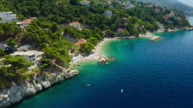 Sahildeki dağ yamacında oteller ve evler. Turistler plajda güneşlenir, denizde yüzer ve kürek çekerler. Hırvatistan 'ın pitoresk Adriyatik kıyısında bir sahil turizm kasabası.