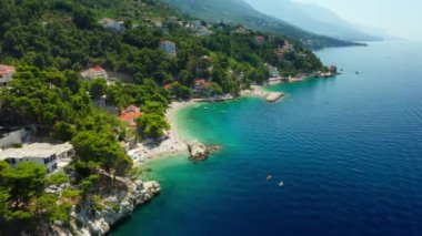 Sahildeki dağ yamacında oteller ve evler. Turistler plajda güneşlenir, denizde yüzer ve kürek çekerler. Hırvatistan 'ın pitoresk Adriyatik kıyısında bir sahil turizm kasabası.