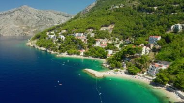 Sahildeki dağ yamacında oteller ve evler. Turistler plajda güneşlenir, denizde yüzer ve kürek çekerler. Hırvatistan 'ın pitoresk Adriyatik kıyısında bir sahil turizm kasabası.