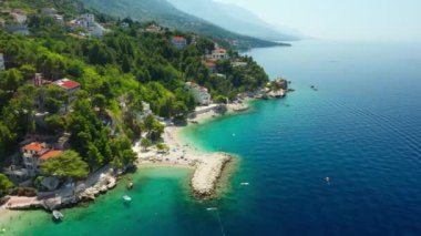 Sahildeki dağ yamacında oteller ve evler. Turistler plajda güneşlenir, denizde yüzer ve kürek çekerler. Hırvatistan 'ın pitoresk Adriyatik kıyısında bir sahil turizm kasabası.