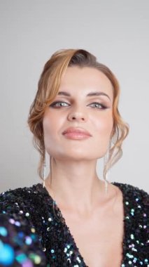 Pullu gece elbisesiyle göz kamaştırıcı genç bir kadın beyaz bir arkaplanda selfie çekiyor. Mükemmel akşam makyajı, yüksek saç stili ve ışıl ışıl küpeler onun büyüleyici görünüşünü tamamlıyor..