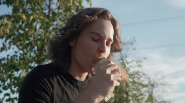 Genç adam açgözlülükle, ambalaj kağıdına sarılı sulu bir parça shawarma 'yı ısırıp çiğniyor. Aç bir adam güneşli bir yaz gününde mavi gökyüzüne karşı sokak yemeği yiyor. Paket servis için doner kebap.