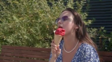 Gevşek saçlı, güneş gözlüklü, inci boncuklu ve yaz elbisesi olan güzel bir kız güneşli bir günde bir bankta oturuyor ve waffle külahında erimiş dondurma yiyor, diliyle yalıyor..