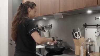 Kadın evde mutfakta yemek pişiriyor. Endüksiyon sobasında. Malzemeleri spatulayla tavada karıştırıyor. Cızırdayan sıcak bir tavada pişene kadar malzemeleri kızartır ve pişirir..