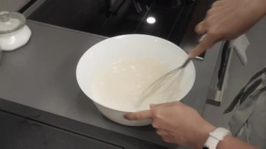 Kadın kasede süt ve yumurta çırpıyor. Ev mutfağında yemek hazırlıyor, hamuru dairesel hareketlerle dikkatlice karıştırıyor. Ev yapımı tarifle krep yapmak için adım adım özel ders.