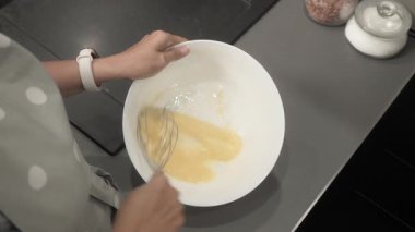 Bir kadın çırpıcıyla bir kasede yumurta çırpıyor. Önlük giyip, mutfağında yemek hazırlıyor ve hamuru hazırlıyor. Ev yapımı tarifle krep yapmak için adım adım özel ders..