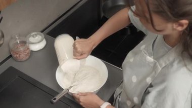 Bir kadın ölçü kabını kullanarak sıvı hamura un döker ve çırpıcıyla karıştırır. Ev mutfağında yemek hazırlıyor, içindekileri bir kasede birleştiriyor. Ev yapımı tarifle krep yapmak..