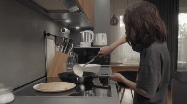 Bir çocuk evde, mutfakta krep kızartır. Bir indüksiyon çadırı üzerinde. Sıvı hamuru sıcak tavaya bir kepçeyle doldurur ve eşit olarak dağıtır. Ev yapımı tarifle krep yapmak..