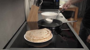 Bir adam evde, mutfakta indüksiyon aletinde krep pişiriyor. Hamuru sıcak tavaya bir kepçeyle doldurur ve eşit olarak dağıtır. Ev yapımı tarifle krep yapmak..