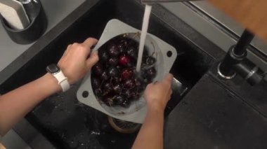 Bir kadın evdeki mutfak lavabosunun üstündeki musluğun altında kiraz yıkıyor. Kocaman böğürtlenleri kevgire akan suyla duruluyor..