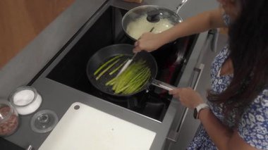 Bir kadın mutfağında kahvaltı için kuşkonmaz hazırlıyor. Yeşil kuşkonmaz mızraklarını kızgın sebze yağında pişiriyor, soba sobasında, onları spatulayla karıştırıyor..