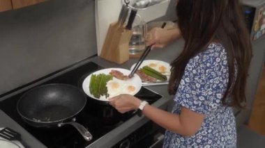 Kahvaltıda kuşkonmaz, yumurta ve pastırma hazırlayan bir kadın. Elinde bir tabak tutuyor ve tavadan tavaya cızırdayan, gevrek domuz pastırması koymak için maşa kullanıyor..