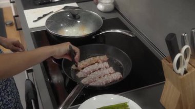 Bir kadın mutfağında indüksiyon sobasında pastırma pişiriyor. Maşa kullanarak, kızgın bir tavada cızırdayan pastırma şeritlerini çeviriyor. Pastırma kendi yağında pişiyor, çıtır çıtır ve altın kahverengiye dönüyor.