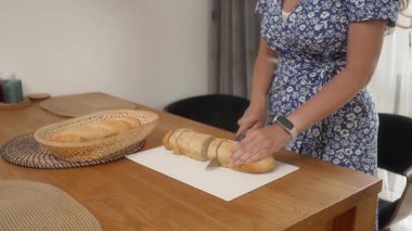 Bir kadın ekmek parçalarını kesme tahtasındaki mutfak bıçağıyla dilimliyor. Onun yanında, ahşap bir mutfak masasında dokunmuş ekmek sepetinde bir tane daha taze ekmek var..