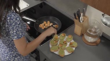 Bir kadın evde kahvaltı için sebze ve deniz ürünleriyle vejetaryen atıştırmalığı hazırlıyor. Avokado soslu sandviçlerin üzerine maşayla kızgın karides koyuyor..