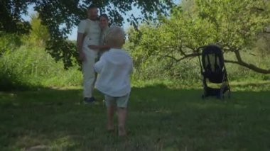 Mutlu aile çifti, çocuklarıyla doğada vakit geçirmekten zevk alır. Bebek sevinçle çimlerin üzerinde ailesine doğru yalınayak koşar. Annem ve babam ağaçların gölgesinde dikilmiş onu izliyorlar..