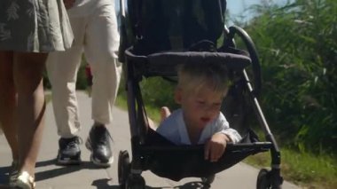 Ebeveynler çocuklarıyla birlikte yürüyorlar. Babam bebek arabasını itiyor. Oyuncak bebek de bebek arabasının içinde, bebek arabasının koltuğunun altında güneşten saklanarak depoya doğru sürünüyor..