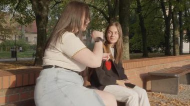 Farklı beden ölçülerinde iki kız arkadaş parkta bir bankta oturuyor, gülümsüyor ve konuşuyor. Biri ince, diğeri büyük bedenli. Kadınlar arasındaki sevgi, arkadaşlık ve derin bağın doğal bir ifadesi..