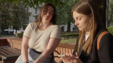 Farklı beden ölçülerinde iki kız arkadaş parkta bir bankta oturuyor, gülümsüyor ve konuşuyor. Biri ince, diğeri büyük bedenli. Kadınlar arasındaki sevgi, arkadaşlık ve derin bağın doğal bir ifadesi..