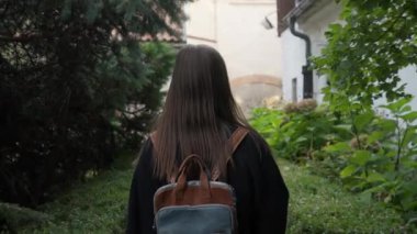 Sırt çantalı genç bir kadın, çalılar ve ağaçlarla çevrili binalar arasında dar yeşil bir ara sokakta yürüyor. Dikiz aynası. Sahne bir yalnızlık, sakinlik ve şehir keşfi hissi veriyor..