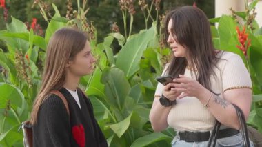 İki kadın dışarıda konuşuyorlar, biri akıllı bir telefon tutuyor ve diğeriyle konuşurken dikkatle dinliyorlar. Etrafları yemyeşil bitkilerle çevrili..