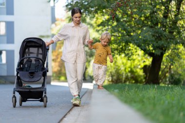 Genç bir anne, parlak bir günde küçük oğluyla parkta yürür. Çocuk onun elini tutuyor, o boş bir bebek arabasını iterken kaldırım kenarında yürüyor. Şehir yeşilliği ve arka plandaki binalar.