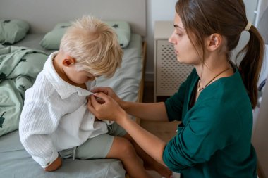 Bir anne, sarışın bebeğinin giyinmesine ve beyaz gömleğinin düğmelerini iliklemesine yardım eder. Sıcak bir yatak odasında sabah rutinlerinde sessiz bir ebeveyn bakımı..
