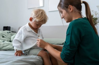 Bir anne, sarışın bebeğine beyaz gömleğiyle yatakta otururken yardım eder. Yatak odasındaki günlük rutinleri sırasında hassas, sessiz bir bakım ve bağlantı anı..