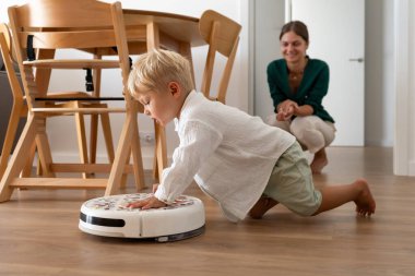 Genç sarışın bir çocuk evdeki ahşap zeminde robot elektrikli süpürgeyle tuhaf bir şekilde etkileşime giriyor. Anne arka planda bir gülümseme ile gözlemler, modern bir aile anı..