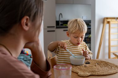 Odaklanmış genç bir çocuk ahşap bir yemek masasında oturur, kaşıkla bir kaseden yemek yer. Yakınlarda bir yetişkin var. Evde aile yemeği sırasında bağımsız yemek yeme anını yakalıyor..