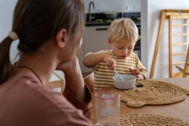 Genç sarışın bir çocuk yemek masasında oturur, kaşıkla bir kaseden yemek yemeye konsantre olur. Bir yetişkin tam karşısında oturur. Sahne, her gün evde sakin bir aile yemeği anı yakalar..