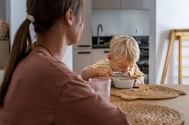 Odaklanmış genç bir çocuk yemek masasında oturur, kaseden yemek için kaşık kullanır. Bir yetişkin tam karşısında oturur. Sahne, günlük aile yemeklerinde bağımsız yeme becerilerini tasvir eder.