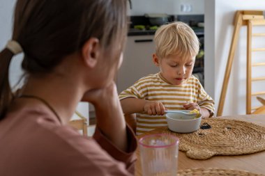Odaklanmış küçük bir çocuk yemek masasında oturur, kaşıkla bir kaseden yemek yer. Yakınlarda yetişkin gözetmenleri var. Olay yeri, evde yemek saatinde bağımsız kendini besleme ve günlük rutin görüntüler yakalıyor.