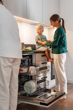 Bir anne küçük çocuğunun ellerini açık bulaşık makinesinin yanındaki mutfak tezgahında çıplak ayakla otururken tutar. Babası yakınlarda duruyor. Aile hayatına ve evdeki paylaşılan anlara odaklan..