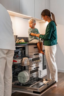 Bir anne küçük çocuğunun ellerini açık bulaşık makinesinin yanındaki mutfak tezgahında çıplak ayakla otururken tutar. Babası yakınlarda duruyor. Ev işleri sırasında aile içi etkileşim sahnesi.