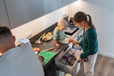 Bir anne ve küçük bir çocuk açık bir mutfak çekmecesine bakarlar, baba ise tezgahın yanında durur. Sahne, günlük aile yaşamını, etkileşimi ve evdeki paylaşılan anları yakalar..